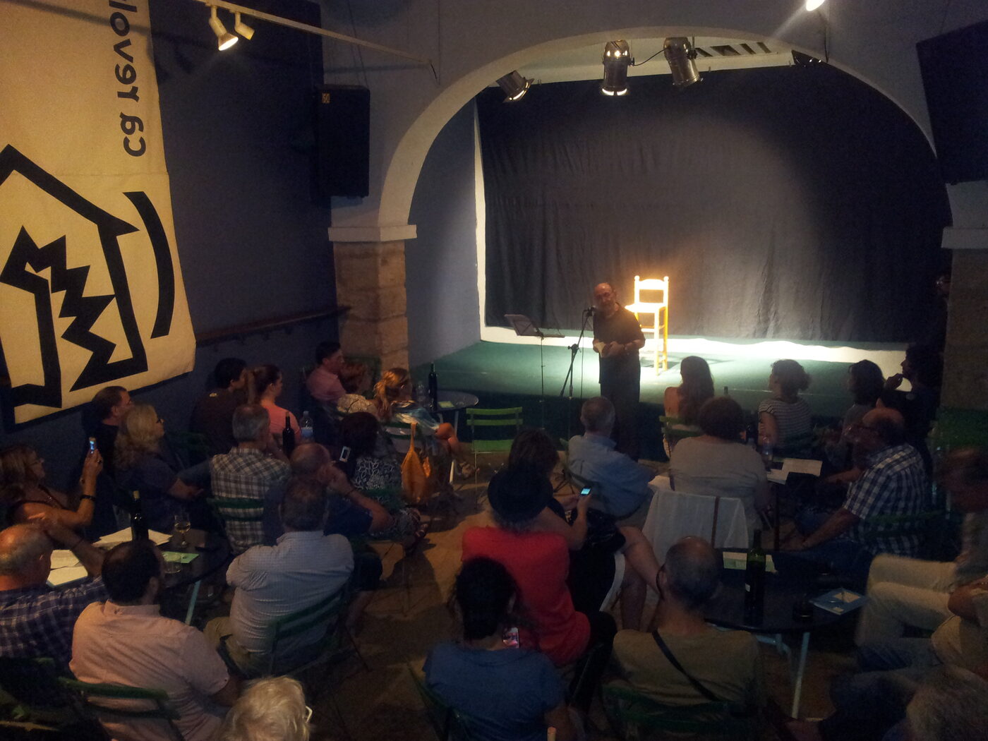 La sala de Ca Revolta, plena de gent asseguda, durant la IV Festa Estellés del 18 de setembre de 2013: al fons, damunt de l'escenari, un home llig davant del micròfon al costat d'una cadira buida.