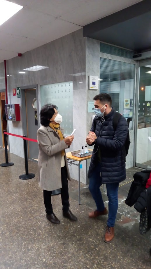 Dues persones amb mascareta conversant al costat d'una taula menuda amb una tauleta digital, al vestíbul d'un edifici amb un cordó roig.