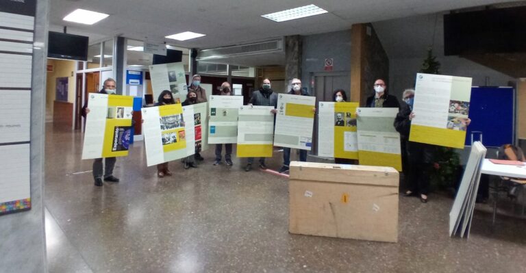 Un grup de persones amb mascareta, dretes al vestíbul d'un edifici, sostenint els panells grocs i blancs de l'exposició sobre l'esperanto.