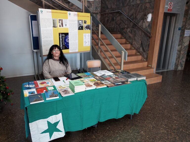 Una dona asseguda darrere d'una taula amb estovalles verdes plena de llibres en esperanto, amb la bandera de l'estel verd davant i, al fons, els panells de l'exposició i una escala.