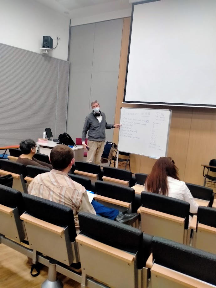 Una persona amb mascareta fent classe davant d'una pissarra blanca amb frases escrites, davant d'unes quantes persones assegudes en una aula.