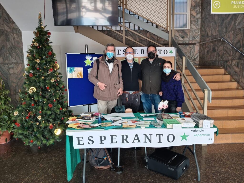 Quatre persones amb mascareta darrere d'una taula informativa amb el rètol «Esperanto» i llibres damunt, al costat d'un arbre de Nadal, al vestíbul d'un edifici.