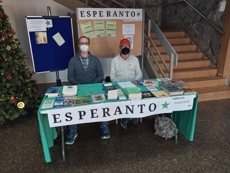 Dues persones amb mascareta assegudes darrere de la taula informativa de l'esperanto, amb llibres damunt i el rètol «ESPERANTO», al costat d'un arbre de Nadal i d'un panell amb la bandera de l'estel verd.
