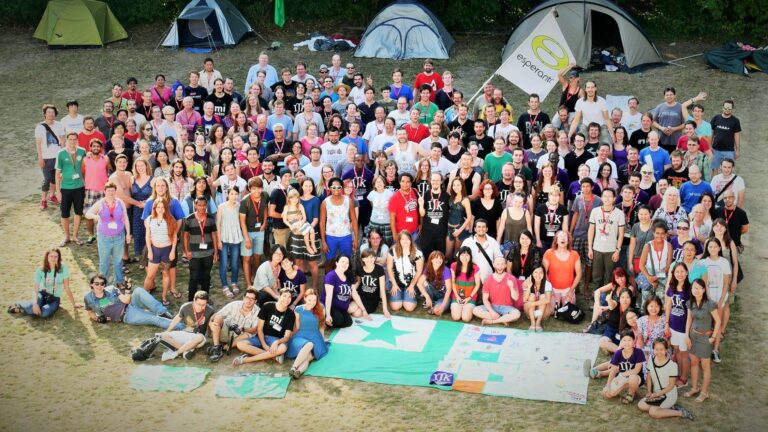 Fotografia des de dalt d'un grup molt nombrós de gent jove posant en una esplanada, en una trobada d'esperanto, amb tendes de campanya al fons, una bandera amb el nom «esperanto» i, damunt de terra, una bandera verda amb l'estel de cinc puntes.