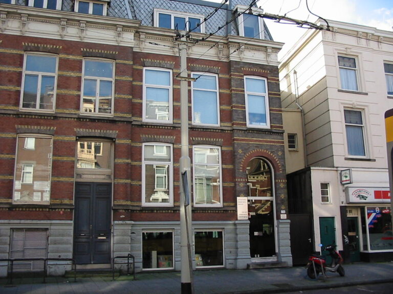 Façana de l'oficina central de la UEA a Rotterdam: una casa de rajola de tres plantes entre mitgeres, amb el número 176 al costat d'una porta d'arc de mig punt, un aparador a peu de carrer amb llibres i un cartell menut amb el nom de l'associació.