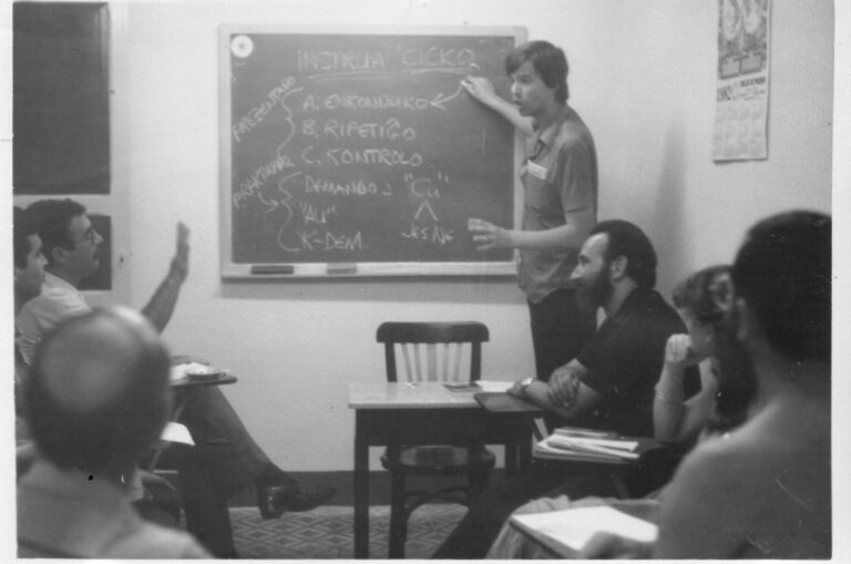 Dennis Keefe dando clase en el grupo; según él, en el local de Fernando el Católico, hacia 1982. Sentado delante, con barba, Pascual Pont. En la pizarra, su «ciclo de enseñanza» (instrua ciklo): introducción, repetición y control; presentación y práctica; preguntas con ĉu, con aŭ y con los interrogativos. Fotografía aportada por Dennis Keefe.