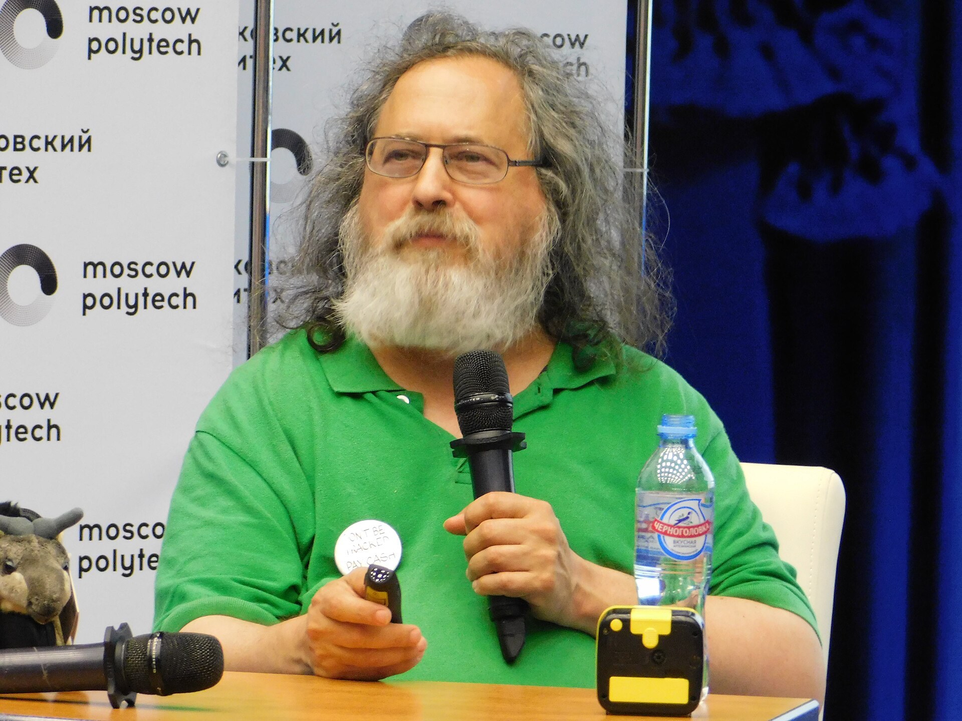 Richard Stallman hablando con un micrófono en la mano, con barba larga y camiseta verde.